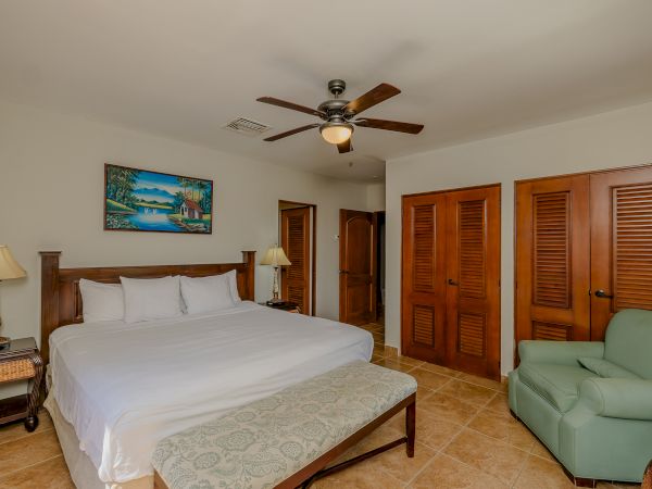 A cozy bedroom features a king-size bed, a green armchair, wooden furniture, a ceiling fan, and a landscape painting on the wall.