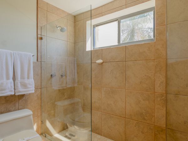 This image shows a modern bathroom with a toilet, glass-walled shower, towel rack, and a window allowing natural light in, all tiled in beige.