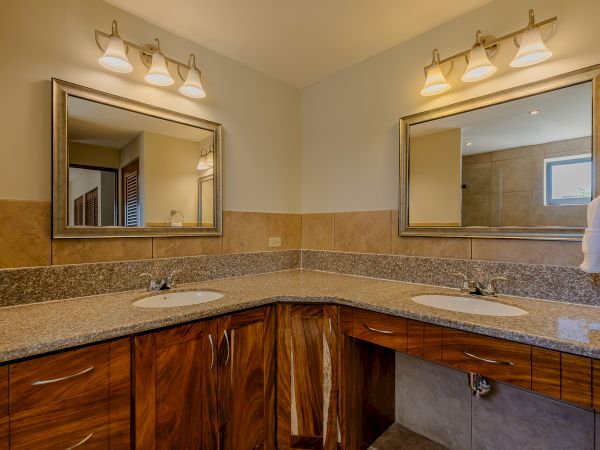 The image shows a modern bathroom with dual sinks, granite countertops, wooden cabinets, and two mirrors, each with three-light fixtures above them.