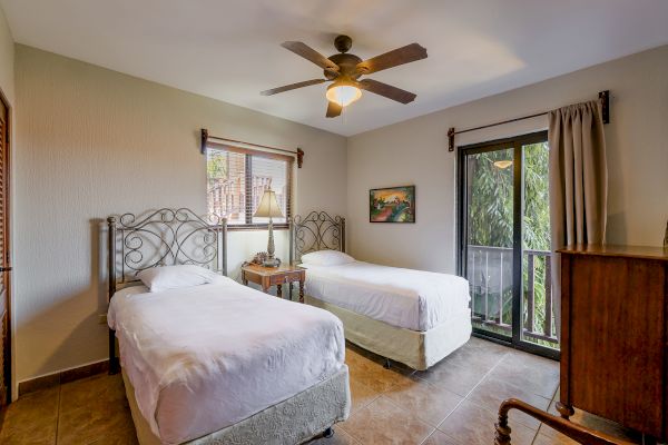 A cozy bedroom with two single beds, a ceiling fan, a nightstand, and large windows covered with curtains allowing natural light into the room.