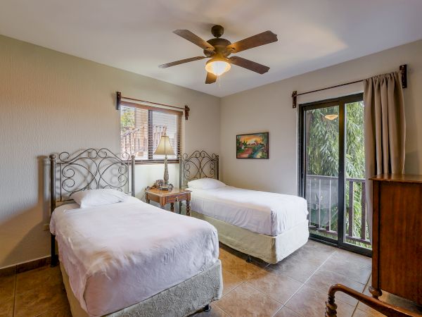A cozy bedroom with two single beds, a ceiling fan, a nightstand, and large windows covered with curtains allowing natural light into the room.