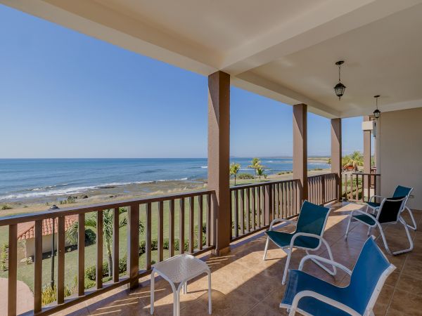 A balcony overlooking a coastal view with several lounge chairs and a small table. The ocean waves can be seen in the distance under a clear sky.