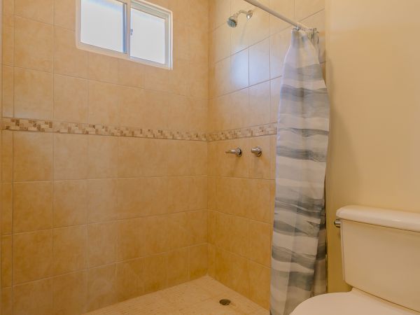 The image shows a bathroom with a toilet, tiled shower area, small window, and a shower curtain. The walls and floors are beige-colored.