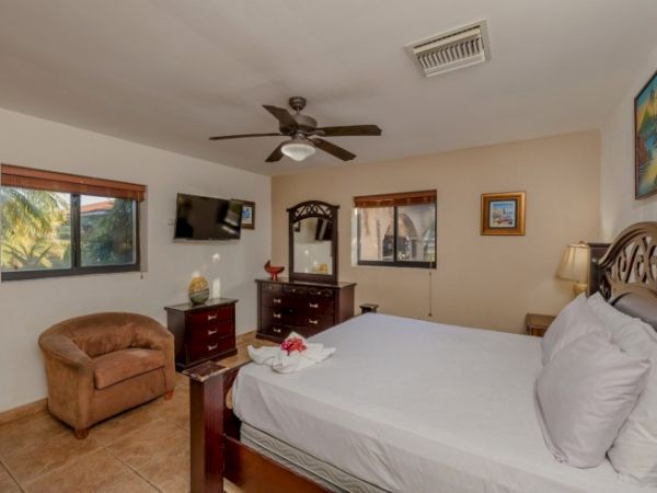 A cozy bedroom with a large bed, dresser, TV, ceiling fan, two windows, an armchair, and decorative artwork on the walls.