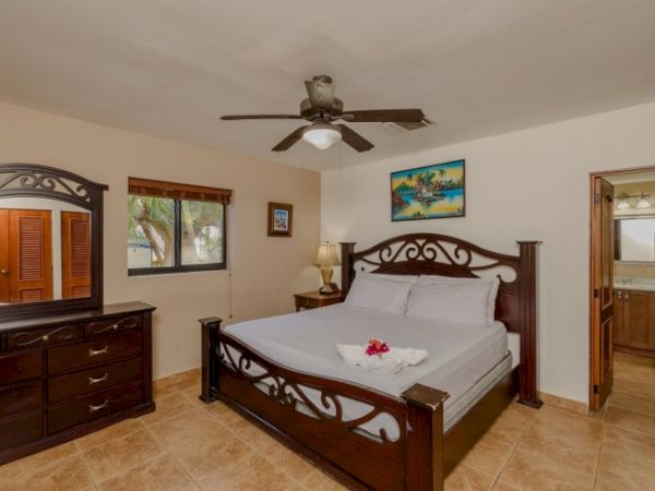 A bedroom features a large wooden bed, dresser, nightstand, ceiling fan, window, and a door leading to a bathroom. Artwork decorates the wall.