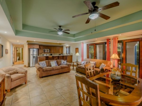 The image shows a spacious living room with two ceiling fans, sofas, a glass dining table, and a kitchen in the background.