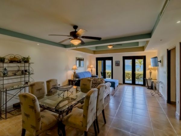 The image depicts a spacious living room with a dining area, sofa, ceiling fan, and large windows with a view of the outdoors.