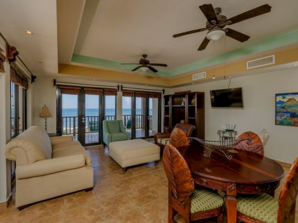 A spacious living room features ceiling fans, large windows with ocean views, two sofas, a round dining table, and a wall-mounted TV ending the sentence.