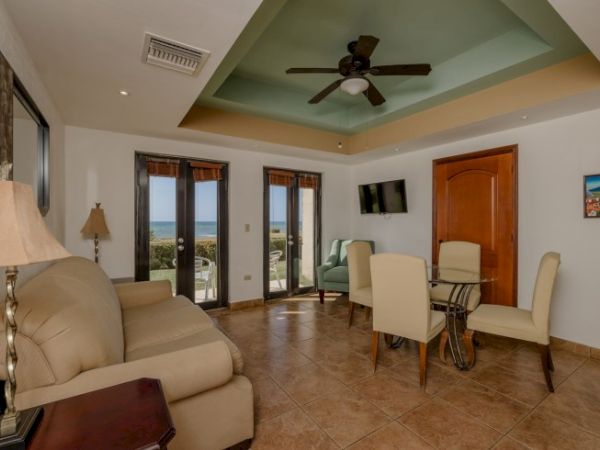 A cozy living room with beige sofas, a wooden table, dining set, ceiling fan, TV, and door. Large windows offer a view of the ocean.