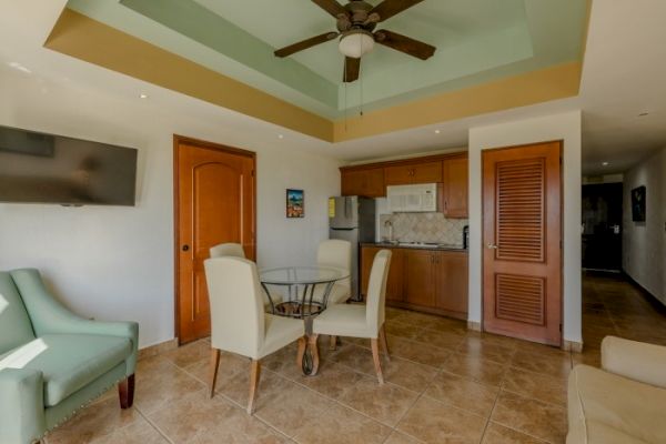 This image shows a room with a ceiling fan, a television, a small dining table with chairs, and a kitchenette area with wooden cabinets and a fridge.