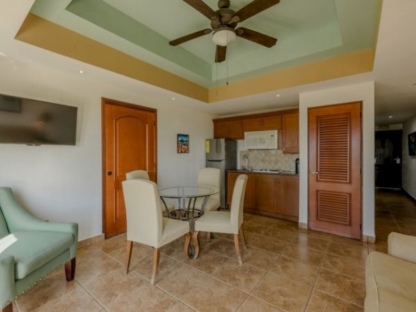 This image shows a room with a ceiling fan, a television, a small dining table with chairs, and a kitchenette area with wooden cabinets and a fridge.
