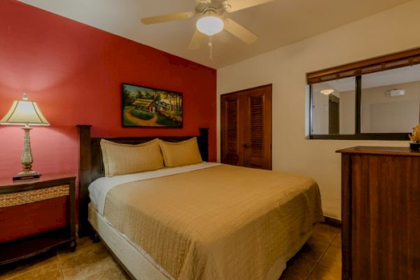 A cozy bedroom with a large bed, a lamp on a bedside table, a painting, and a ceiling fan. There's a dresser and a window with wooden blinds.