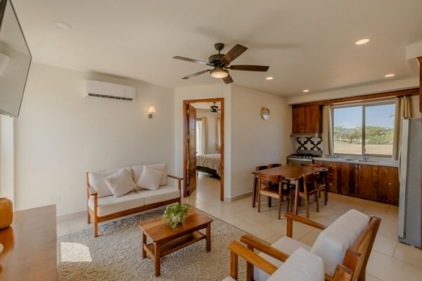 The image shows a cozy living room with a ceiling fan, a small kitchen, and a dining area. There's a sofa set, coffee table, and window.