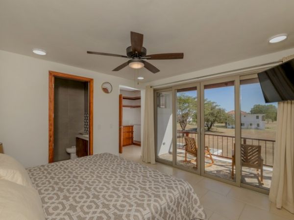 A bedroom with a ceiling fan, patterned bedspread, wall-mounted TV, en-suite bathroom, large sliding glass doors, and a balcony with outdoor seating.
