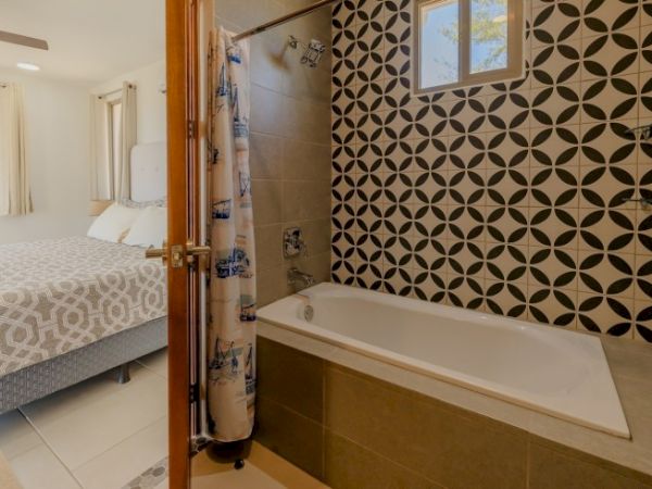 The image shows a bathroom with a bathtub and patterned tiles on the wall. An open door reveals an adjoining bedroom with a bed and matching linens.