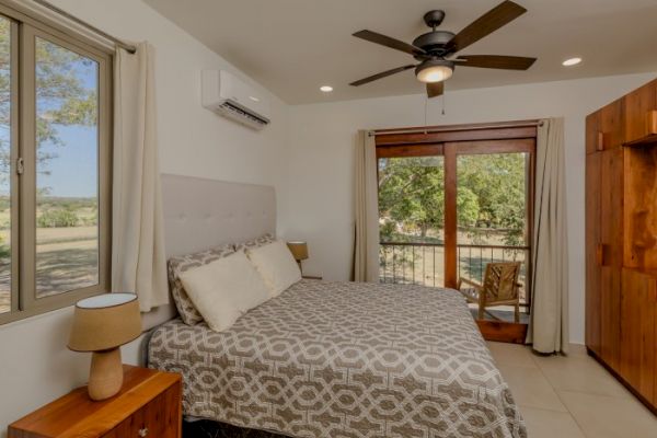 The image shows a cozy bedroom with a double bed, large window, ceiling fan, wooden furniture, and balcony view of greenery.