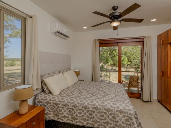 The image shows a cozy bedroom with a double bed, large window, ceiling fan, wooden furniture, and balcony view of greenery.