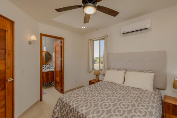 A bedroom with a bed, nightstands, a ceiling fan, and an air conditioner. The door leads to a bathroom with a visible sink and mirror.