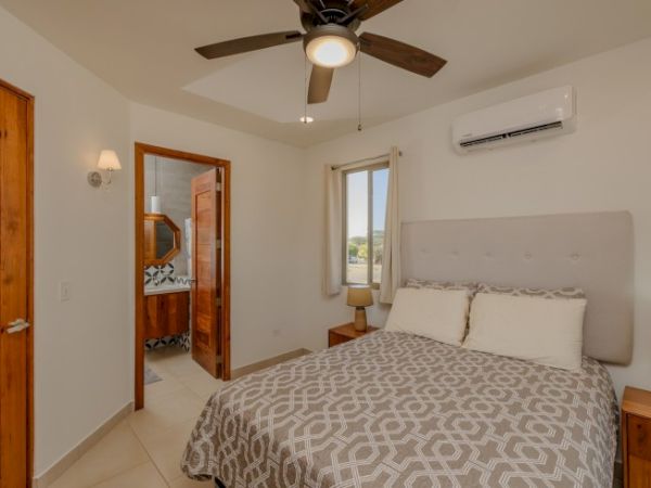 A bedroom with a bed, nightstands, a ceiling fan, and an air conditioner. The door leads to a bathroom with a visible sink and mirror.