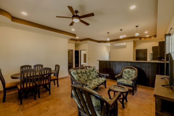 The image shows a cozy living room with a dining table, ceiling fan, cushioned chairs, a TV, and a kitchen area with a counter and hanging lights.