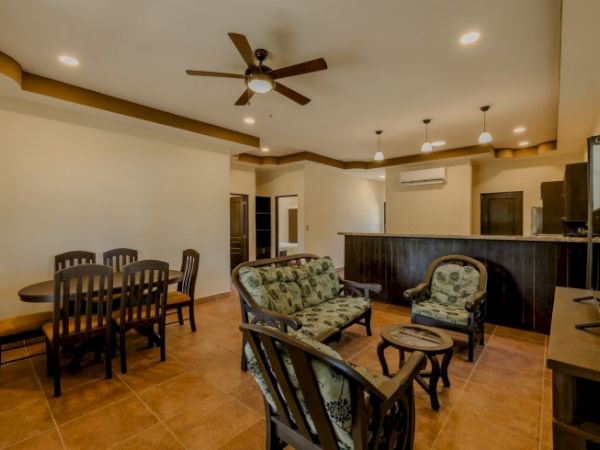 The image shows a cozy living room with a dining table, ceiling fan, cushioned chairs, a TV, and a kitchen area with a counter and hanging lights.