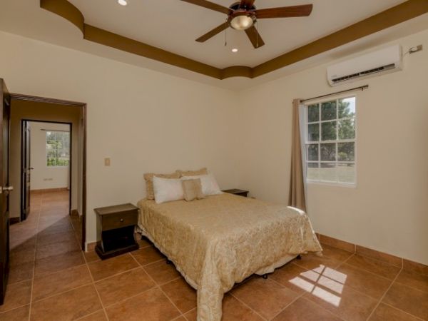 A bedroom with a ceiling fan, air conditioner, bed with beige bedding, and two bedside tables. An open door shows a hallway and another room.