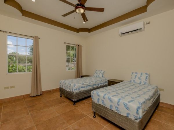 A room with two twin beds, patterned bedding, ceiling fan, air conditioner, and two windows with curtains. Light tiled floor with wooden bed frames.