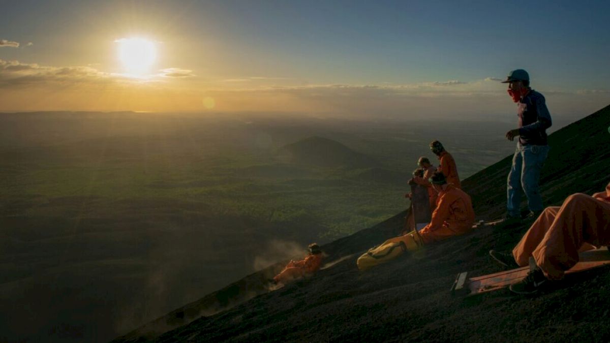 People in orange suits are sitting and standing on a steep, rocky slope with an expansive, green landscape in the background, at sunset.