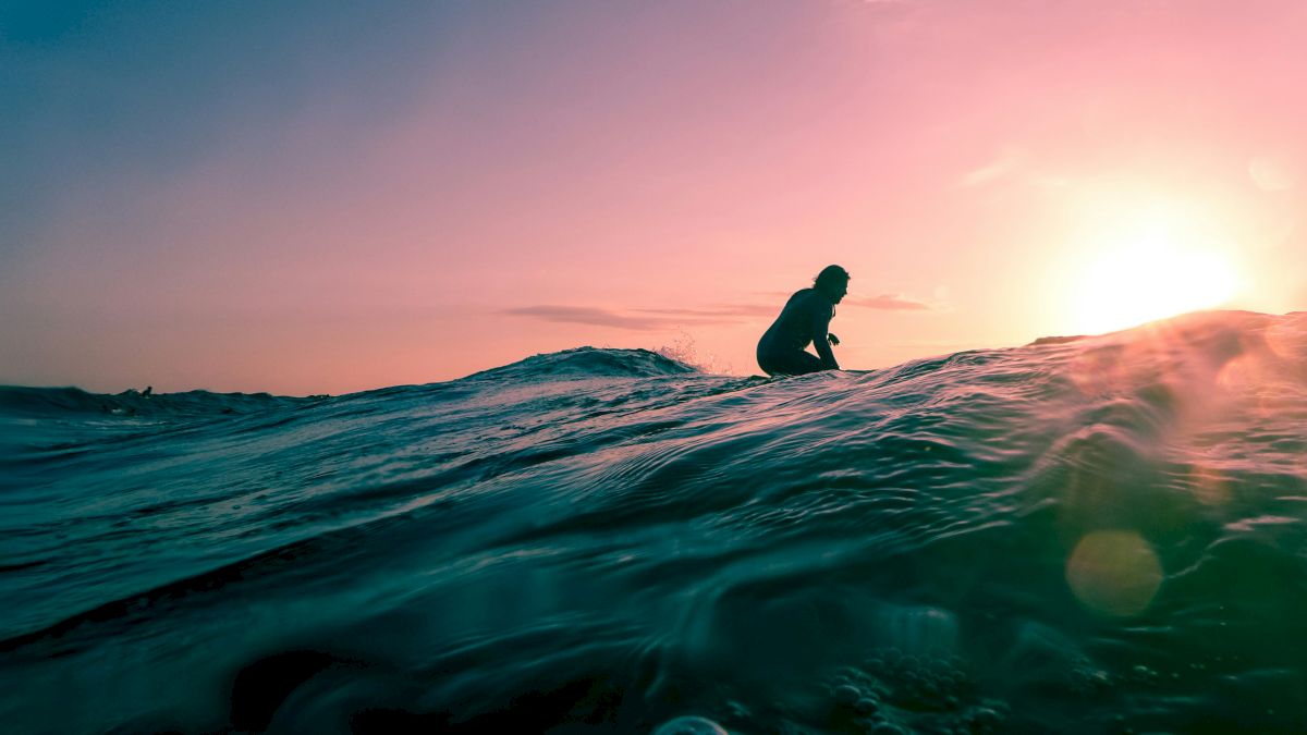 A silhouette of a person is seen surfing on a wave during a sunset, captured from the water's surface, with a vibrant sky in the background.