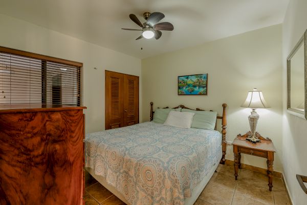 A cozy bedroom with a ceiling fan, a double bed with patterned cover, bedside table with lamp, dresser, window with blinds, and framed artwork.