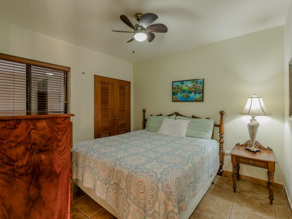 A cozy bedroom with a ceiling fan, a double bed with patterned cover, bedside table with lamp, dresser, window with blinds, and framed artwork.