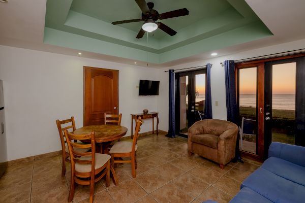 The image shows a living room with a dining table and chairs, a sofa, an armchair, a ceiling fan, a TV, and a view of a sunset through glass doors.