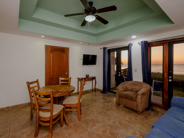 The image shows a living room with a dining table and chairs, a sofa, an armchair, a ceiling fan, a TV, and a view of a sunset through glass doors.