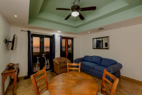 A cozy living room with a round dining table, chairs, blue couch, armchair, TV, ceiling fan, balcony with sunset view, and wall mirror ends the sentence.