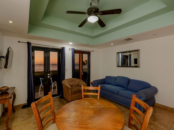 A cozy living room with a round dining table, chairs, blue couch, armchair, TV, ceiling fan, balcony with sunset view, and wall mirror ends the sentence.