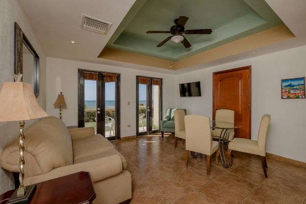 A living room with a beige sofa, chairs, a ceiling fan, a wall-mounted TV, and large windows offering an ocean view, alongside a small dining area.