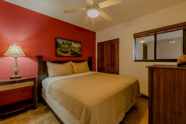 This image shows a bedroom with a double bed, a red accent wall, a bedside table with a lamp, a painting above the bed, and a ceiling fan.