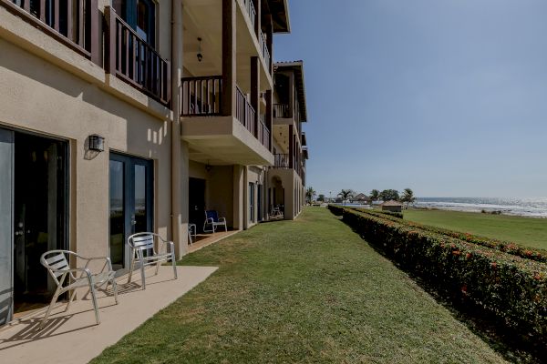 The image shows an apartment building with balconies and a patio area, overlooking a grassy area and the ocean on a clear day.