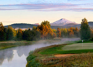 A picturesque scene of a golf course by a river with mist rising, surrounded by trees and a snow-capped mountain in the background at sunrise.