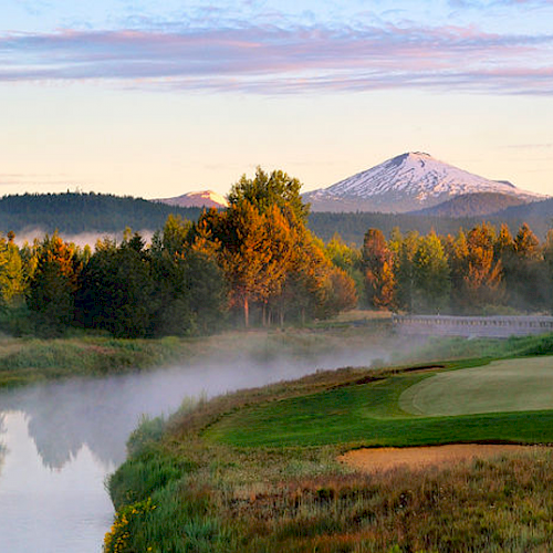 A picturesque scene of a golf course by a river with mist rising, surrounded by trees and a snow-capped mountain in the background at sunrise.