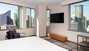A modern hotel room with large windows offering a cityscape view, featuring a bed, armchair, TV, wooden bench, and small side table.