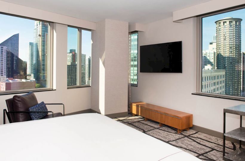 A modern hotel room with large windows offering a cityscape view, featuring a bed, armchair, TV, wooden bench, and small side table.