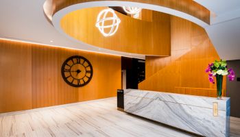 The image shows a modern lobby with a marble reception desk, a large wall clock, and a curved wooden structure with hanging spherical lights.
