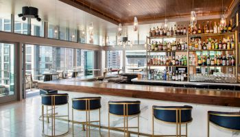 A modern bar with a wooden top, elegant high chairs, and a well-stocked assortment of bottles on shelves in a bright setting with city views.