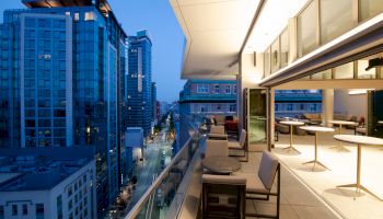 A modern cityscape view from a balcony at dusk, featuring tall buildings, streetlights, tables, and chairs on the balcony.