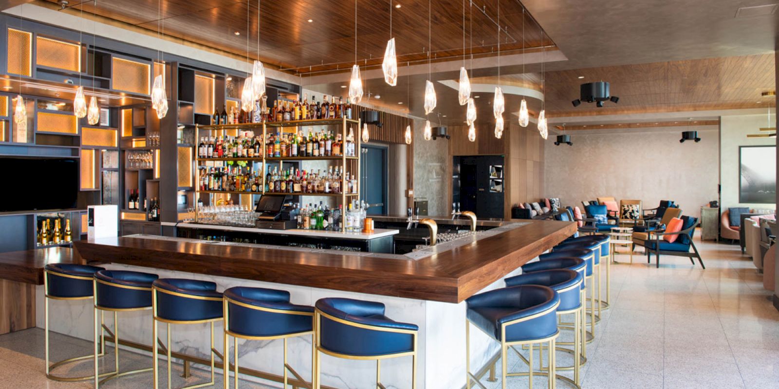 This image shows a modern bar with a long counter, blue cushioned stools, hanging lights, a well-stocked bar, and a lounge area in the background.