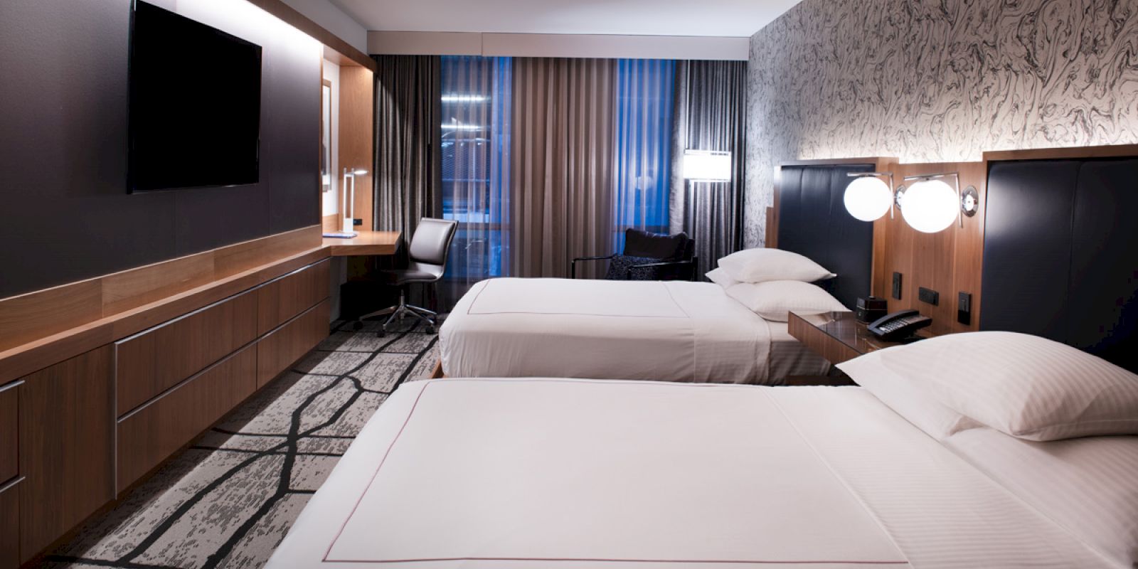 The image shows a modern hotel room with two beds, a large TV, a work desk, and contemporary decor, featuring a patterned wall and large windows.