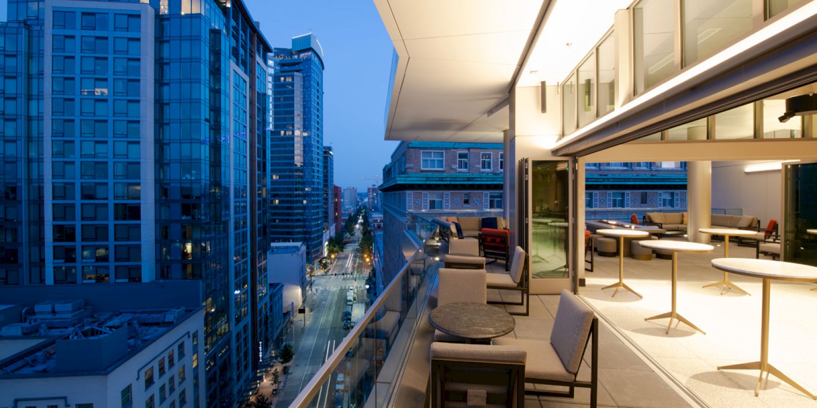A modern balcony with tables and chairs overlooks a city street lined with tall buildings at dusk, featuring expansive glass windows and a cozy atmosphere.