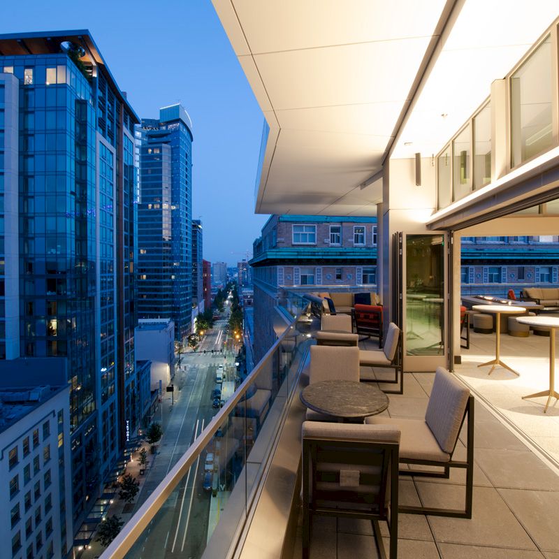 A balcony with seating areas overlooks a cityscape at dusk, featuring modern buildings illuminated against a twilight sky.