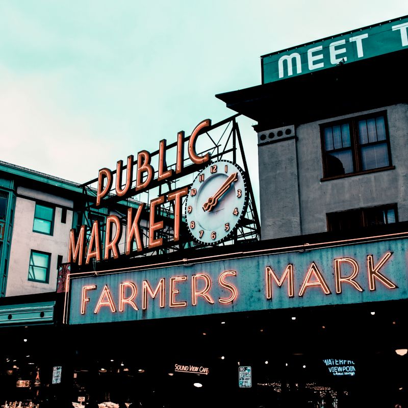 The image shows the iconic neon sign for a public farmers market in an urban setting, with additional surrounding buildings visible.